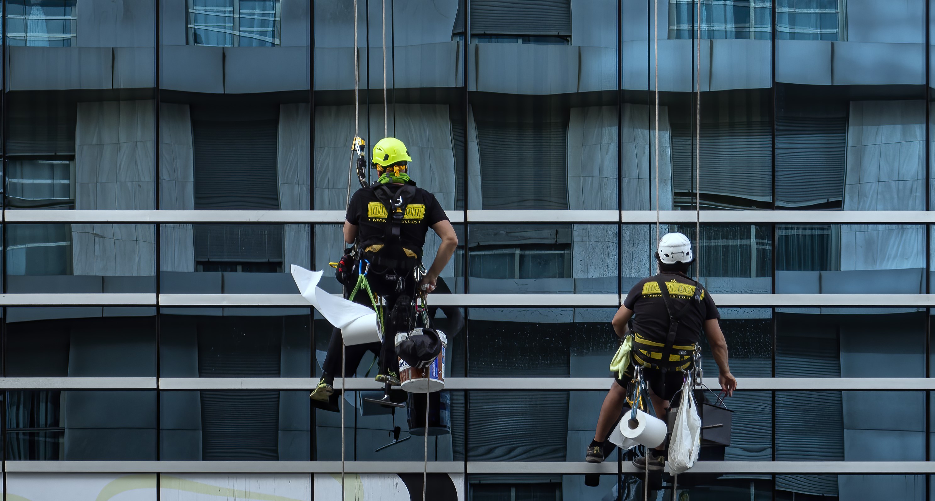 En la fotografía de esta noticia se pueden ver dos hombres en un andamio vertical limpiando las ventanas de un edificio.