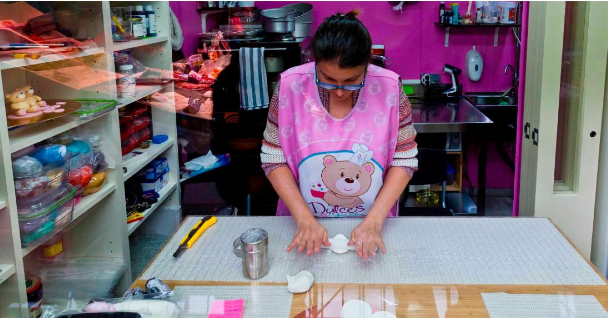 Mujer vestida con un delantal rosa y un oso de peluche elaborando dulces en un taller tipo obrador. 