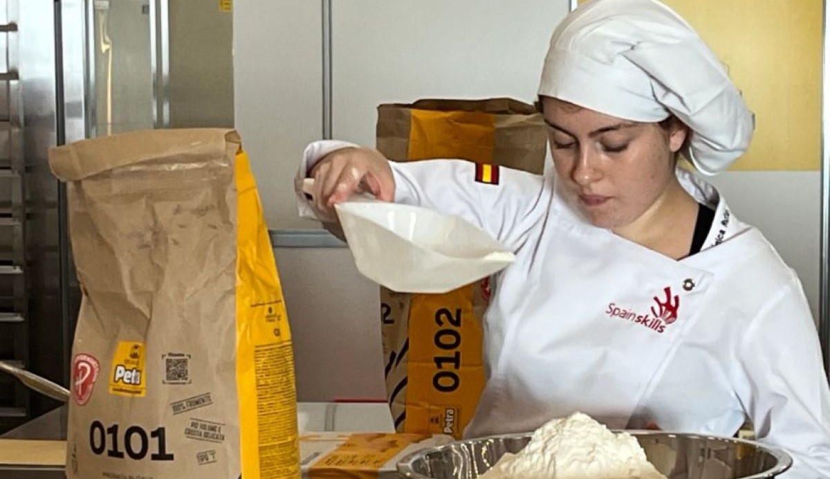 Mujer elaborando algún tipo de alimento añadiendo harina a un bol. Va vestida con uniforme de cocina.