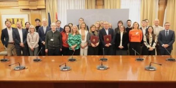 Fotografía de la firma del Convenio. Un gran número de personas detrás de una mesa de madera repleta de micrófonos. Dos de las personas más centradas portan unas carpetas.